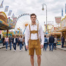 A man stands in the center of a fairground wearing traditional Bavarian attire: a white long-sleeve shirt with a tan collar, brown leather-like overalls with gold embroidery, and matching brown lederhosen. 