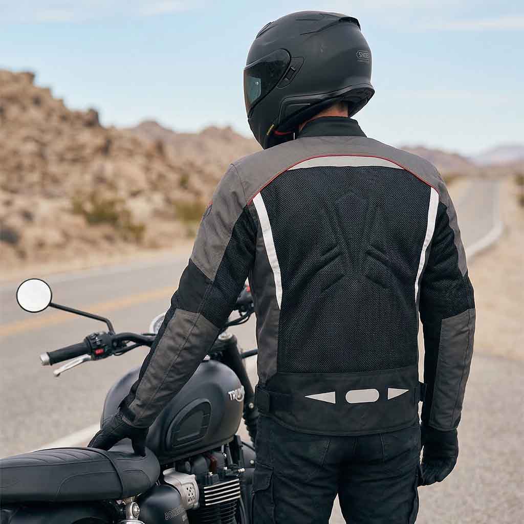 Person wearing a black and gray motorcycle jacket and helmet standing next to a motorcycle on a desert road.