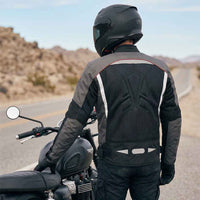 Person wearing a black and gray motorcycle jacket and helmet standing next to a motorcycle on a desert road.