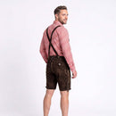 A man wearing traditional Bavarian-style dark brown leather shorts with suspenders, a red and white checkered long-sleeve shirt, and dark suspenders, photographed against a plain white studio background.
