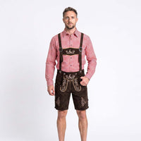 A man modeling traditional Bavarian attire consisting of dark brown suede lederhosen with embroidered details and suspenders, paired with a red and white gingham long-sleeve shirt, against a plain white studio background.